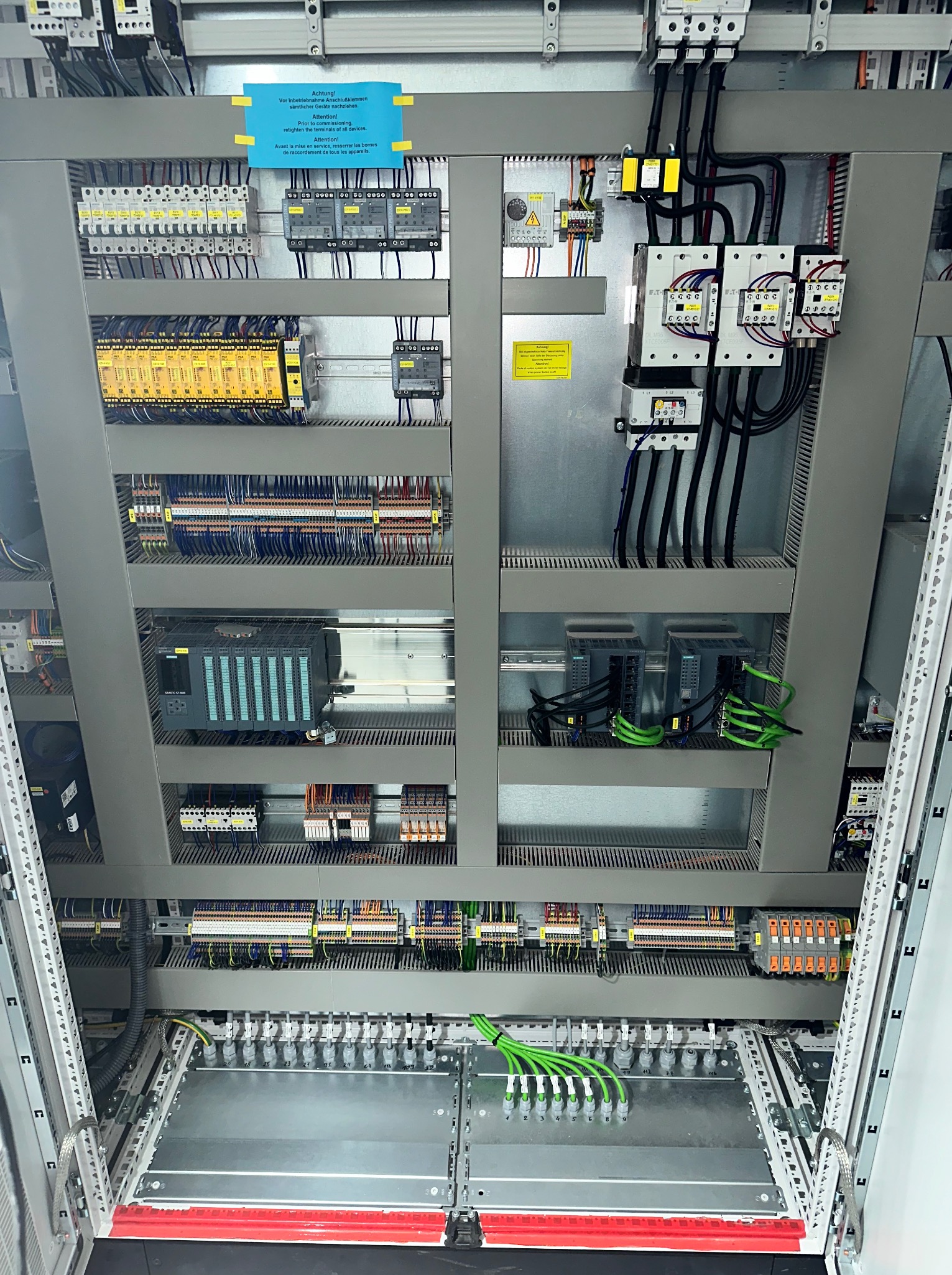 Wnętrze rozdzielnicy elektrycznej z licznymi przewodami, modułami i zabezpieczeniami. Widoczne listwy zaciskowe, sterowniki PLC oraz przewody w kolorach czarnym i zielonym.