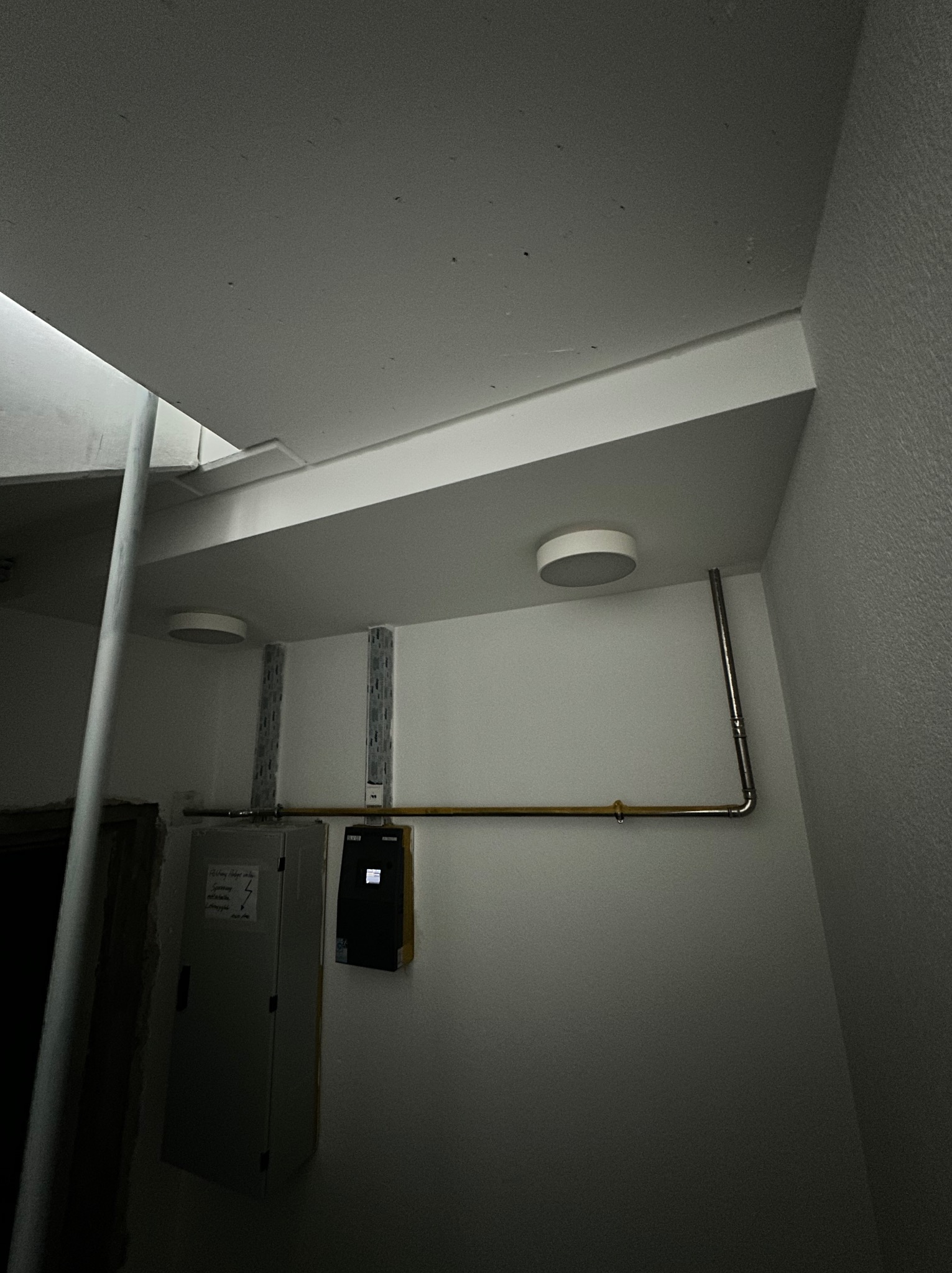Szare wnętrze z zamontowanym oświetleniem sufitowym, widoczne także rury i skrzynka elektryczna na ścianie. Ujęcie z dołu, ciemne otoczenie.