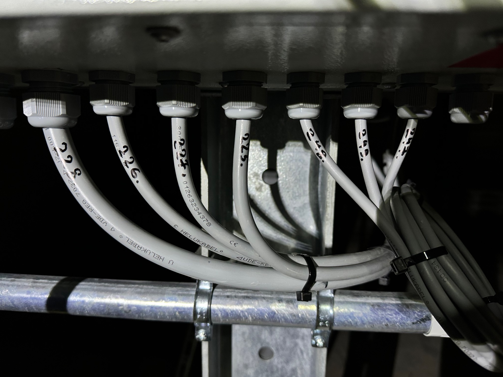 Szereg kabli wchodzących do skrzynki przyłączeniowej, zabezpieczonych dławikami, przymocowanych do metalowej konstrukcji. Widoczne numery identyfikacyjne na kablach.