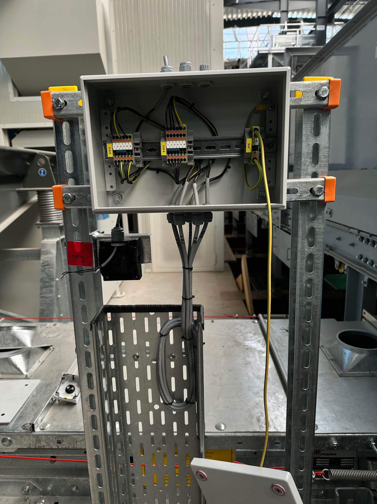 Otwarta skrzynka elektryczna z podłączonymi przewodami, zamontowana na metalowej konstrukcji w hali przemysłowej. Widoczne złącza, listwy zaciskowe i kable w różnych kolorach.