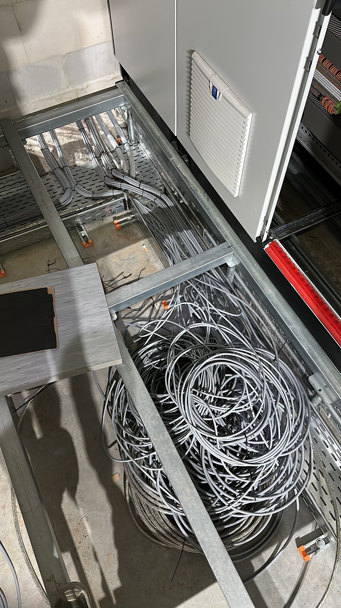 Kable elektryczne ułożone w metalowych korytach, widoczne elementy rozdzielnicy, szare przewody w zwojach i uporządkowane odcinki, instalacja w trakcie realizacji.