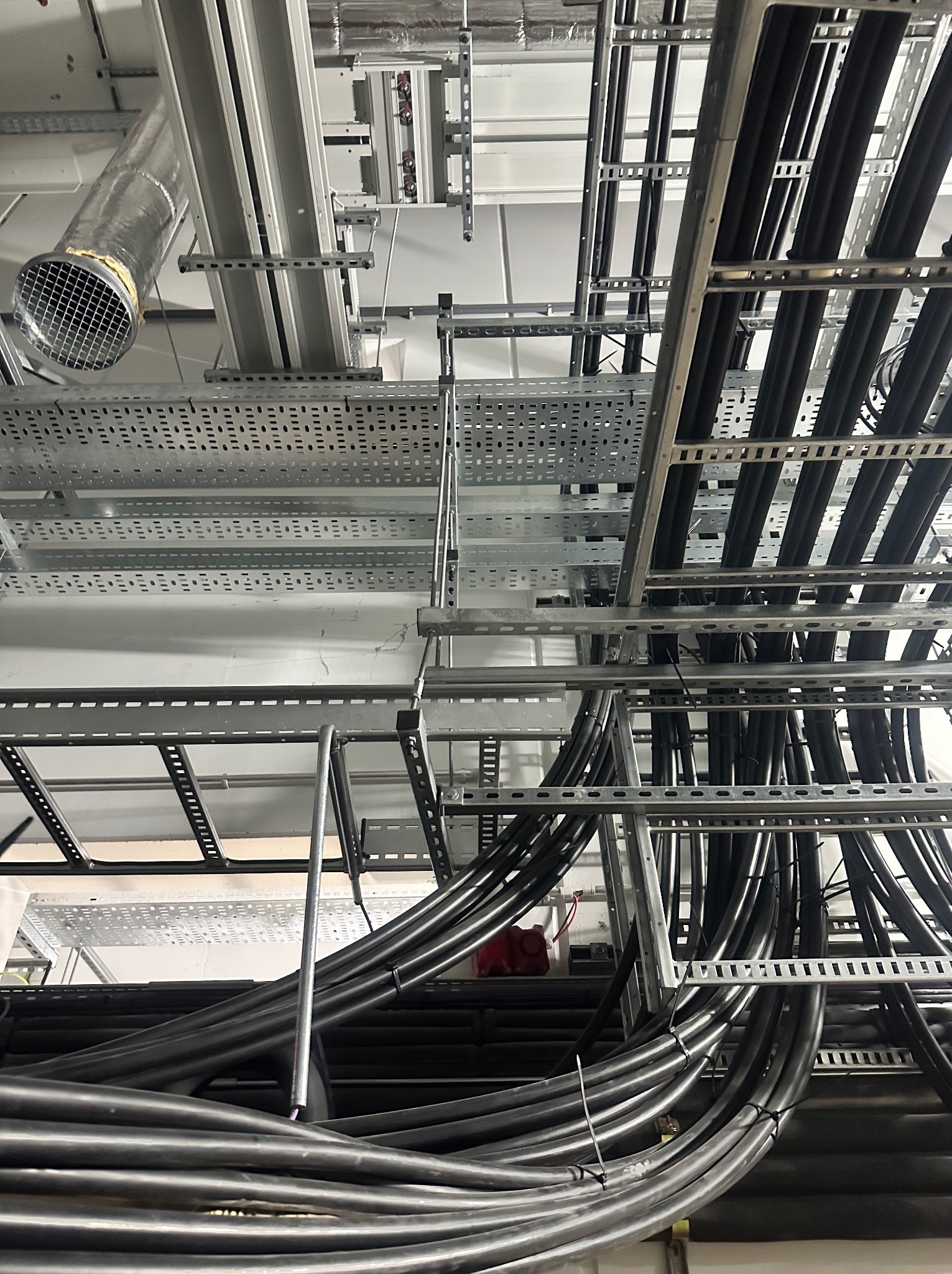 Widok z dołu na koryta kablowe z przewodami w budynku komercyjnym. Metalowe konstrukcje pod sufitem, wentylacja, przewody ułożone równolegle.