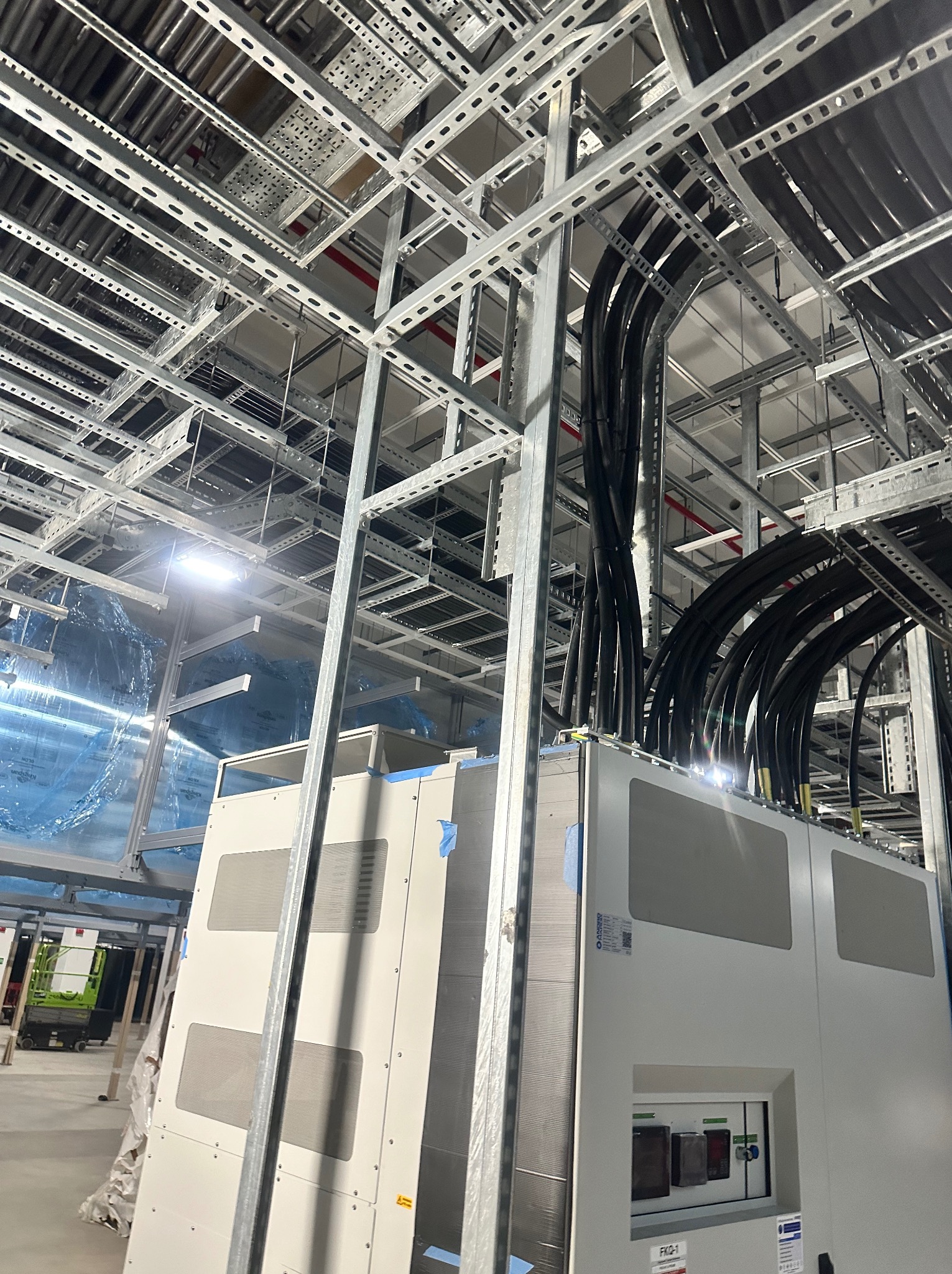 Kable elektryczne prowadzone w metalowych korytach pod sufitem w nowym budynku. Widoczne szafy elektryczne i elementy konstrukcyjne sufitu.