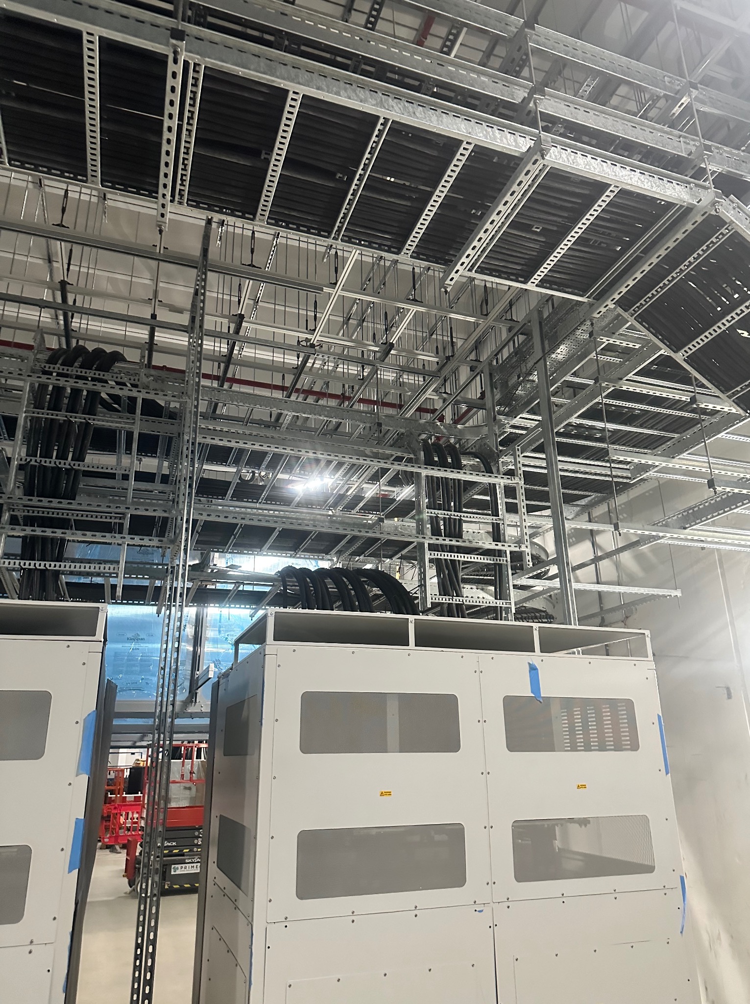 Metalowe koryta kablowe pod sufitem w hali przemysłowej, widoczne białe szafy elektryczne i podnośnik koszowy w tle.