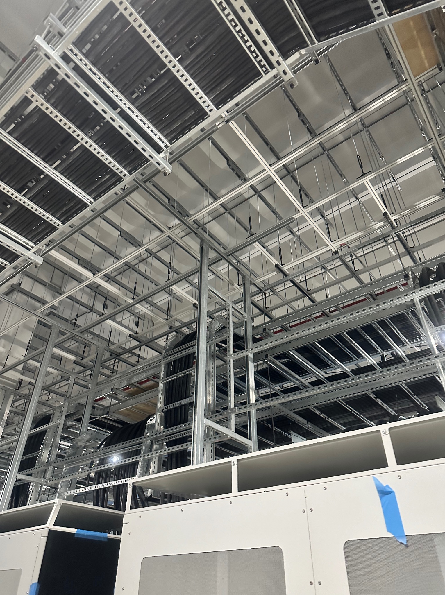 Skomplikowana, metalowa konstrukcja pod sufitem z licznymi kablami. Widoczne białe szafy z niebieską taśmą. Perspektywa z dołu ukazuje złożoność instalacji.