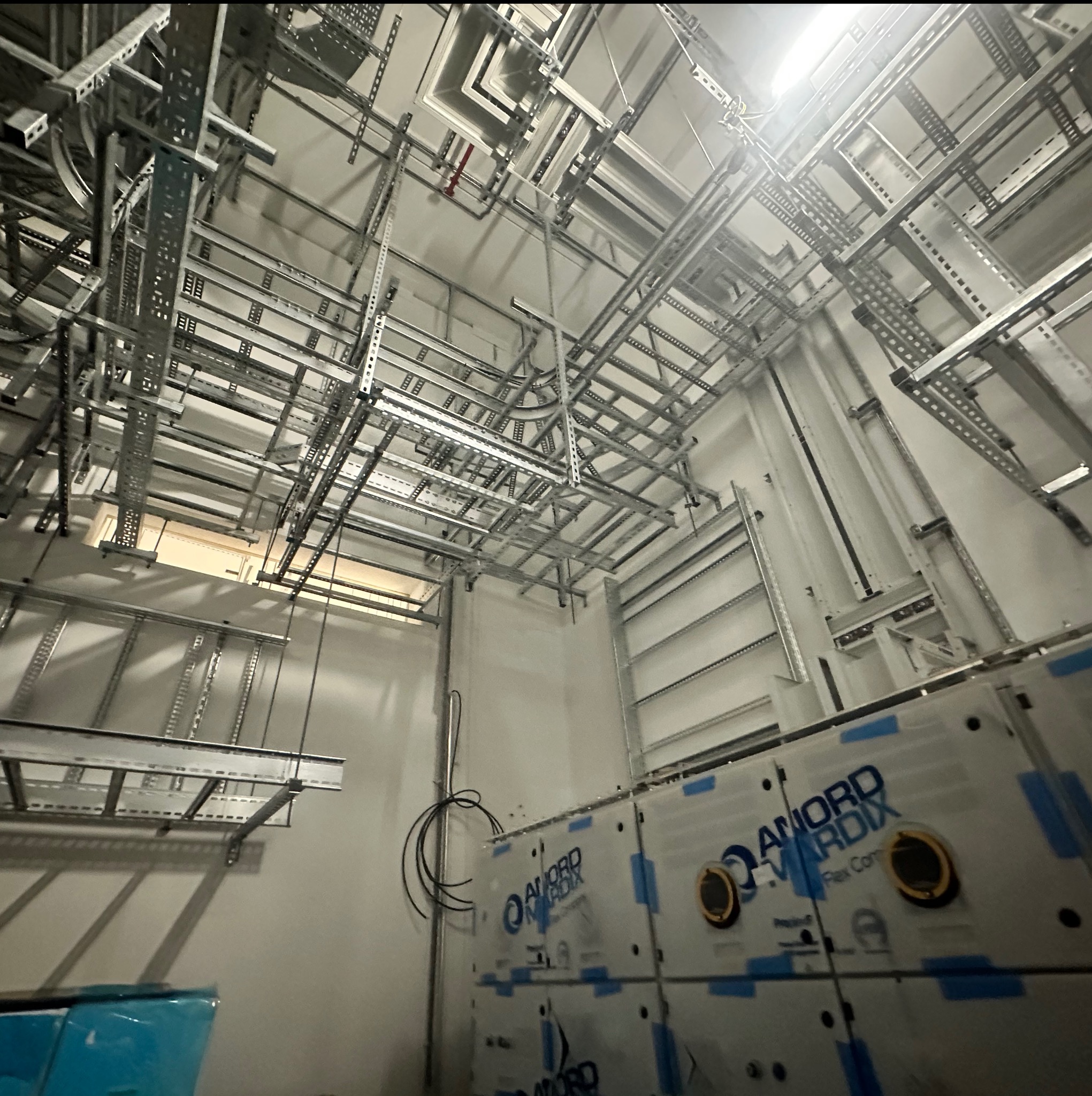 Skomplikowana sieć metalowych korytek kablowych i wsporników pod sufitem w jasnym pomieszczeniu. Widoczne również kable i urządzenia.