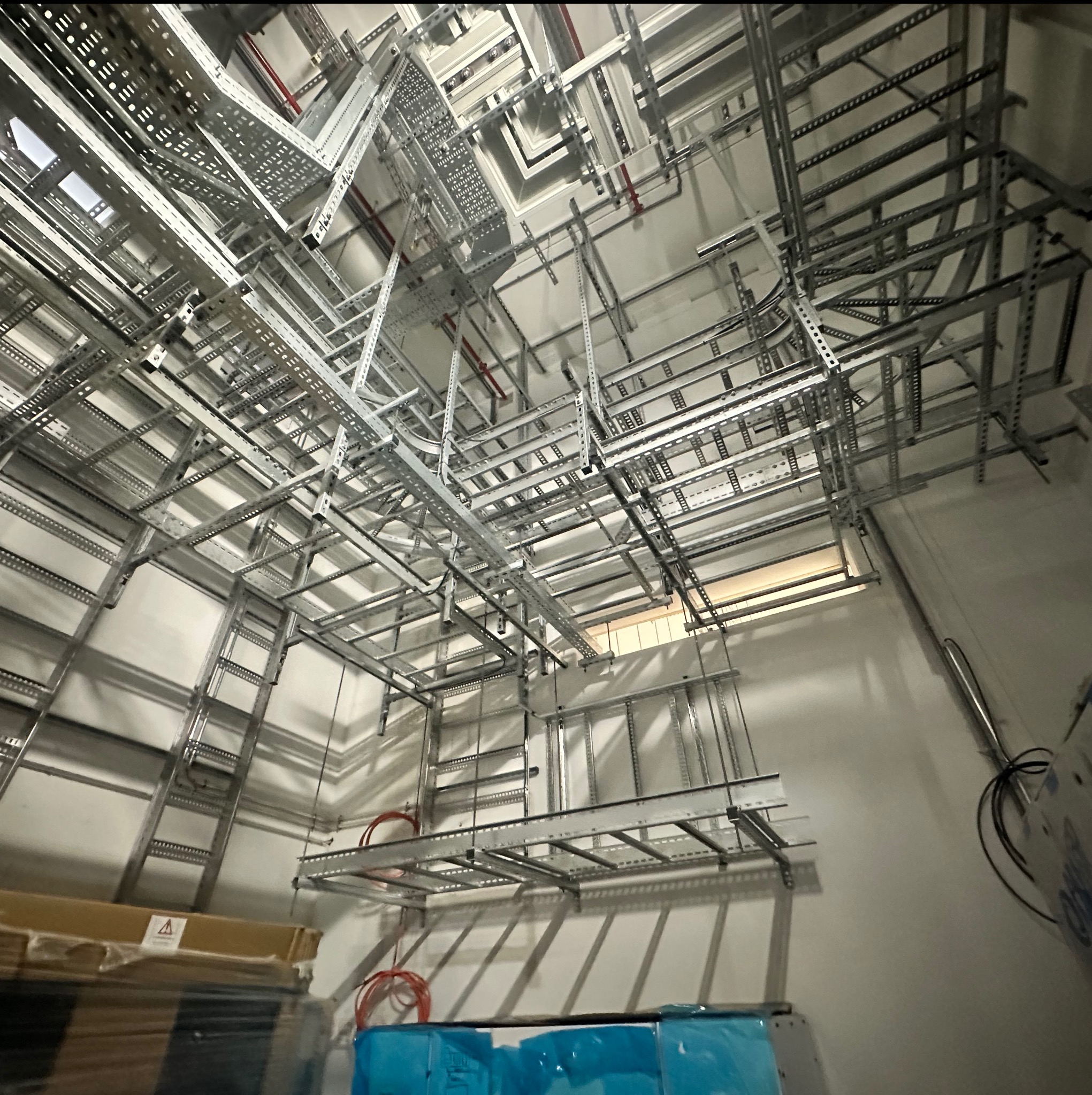 Skomplikowana sieć metalowych korytek kablowych pod sufitem, częściowo z przewodami. Widok z dołu, oświetlenie mieszane.