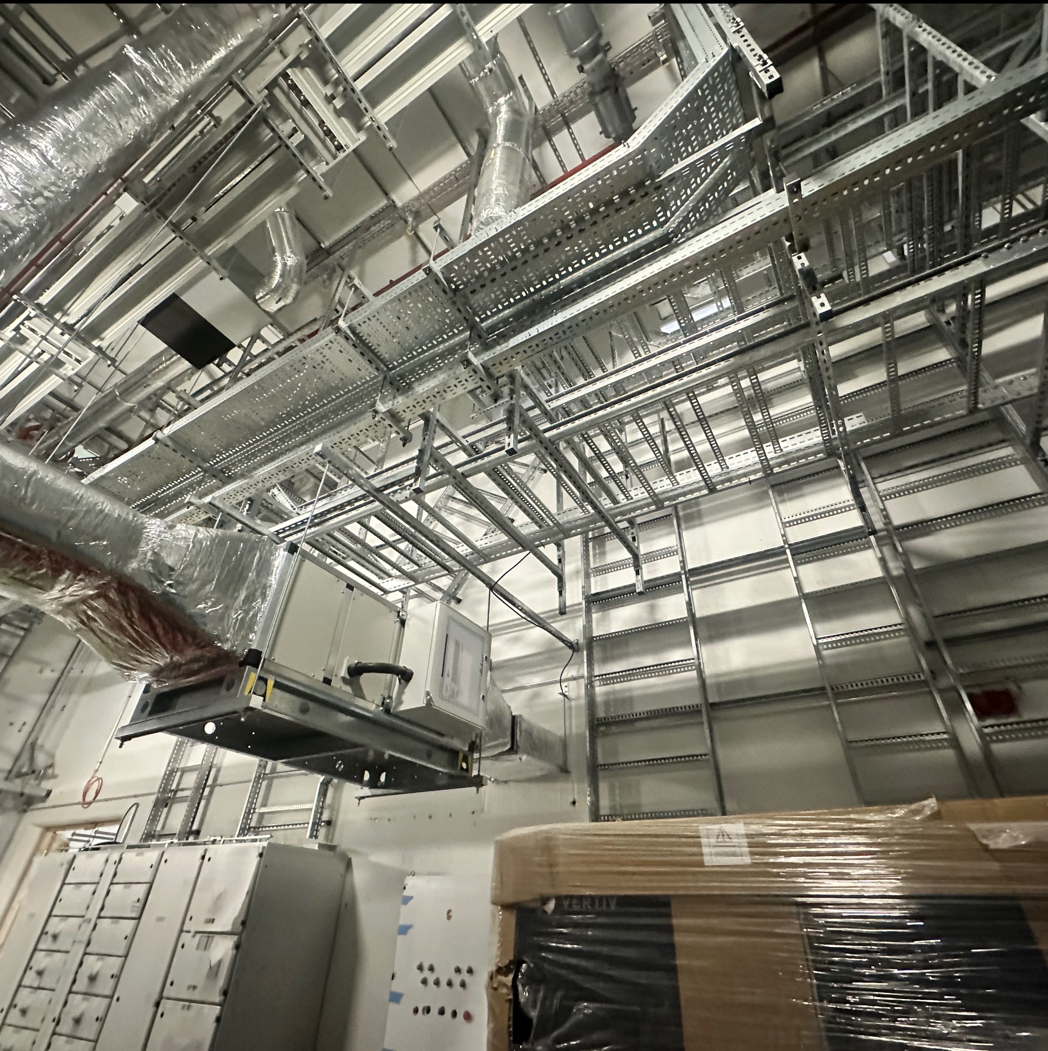 Skomplikowana sieć metalowych korytek kablowych i rur wentylacyjnych pod sufitem hali. Widoczne szafy sterownicze i zapakowane urządzenia.