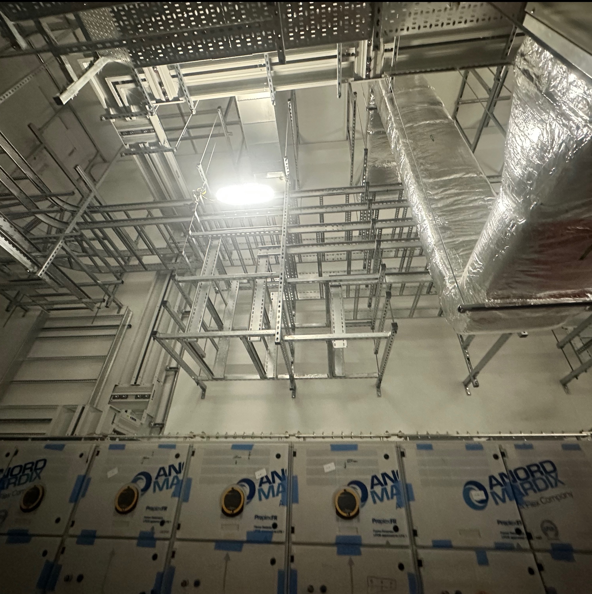 Metalowe koryta kablowe pod sufitem w hali przemysłowej, widoczne kanały wentylacyjne w srebrnej izolacji. Instalacje elektryczne w trakcie montażu.