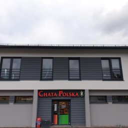 Fach-Mal - Dwukondygnacyjny budynek z szarą elewacją i ciemnoszarymi panelami. Nad wejściem czerwony napis 'CHATA POLSKA'. Widok na fasadę budynku w pochmurny dzień.