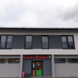Dwukondygnacyjny budynek z szarą elewacją i ciemnoszarymi panelami. Nad wejściem czerwony napis 'CHATA POLSKA'. Widok na fasadę budynku w pochmurny dzień.