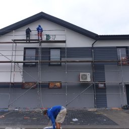 Fach-Mal - Dwóch pracowników na rusztowaniu maluje dwukolorową elewację budynku. Widoczny klimatyzator na ścianie i osoba przygotowująca teren do malowania.