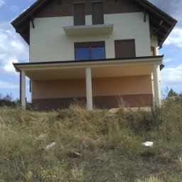 Fach-Mal - Dwukondygnacyjny dom z beżową elewacją i drewnianym dachem, widziany z dołu z zarośniętego wzgórza. Widoczne kolumny podtrzymujące balkon i brązowe okiennice.