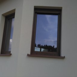 Fach-Mal - Elewacja domu po malowaniu w jasnym odcieniu, z brązowymi ramami okiennymi. Widoczne dwa okna z odbiciem nieba i drzew. Ujęcie z dołu, ukazujące fragment dachu.