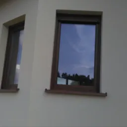 Elewacja domu po malowaniu w jasnym odcieniu, z brązowymi ramami okiennymi. Widoczne dwa okna z odbiciem nieba i drzew. Ujęcie z dołu, ukazujące fragment dachu.