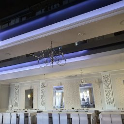 Fach-Mal - Elegancka sala bankietowa z dekoracyjnymi panelami ściennymi, lustrami i kryształowymi żyrandolami. Sufit podwieszany z niebieskim oświetleniem LED. Białe krzesła i stoły.