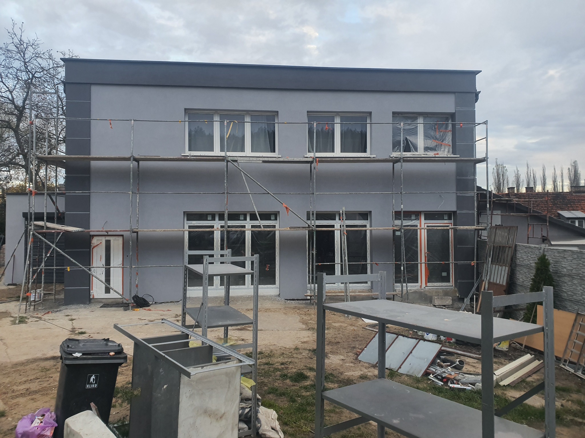 Szara elewacja dwukondygnacyjnego domu w trakcie malowania, z rusztowaniami i widocznymi elementami przygotowawczymi. Ujęcie z poziomu gruntu.