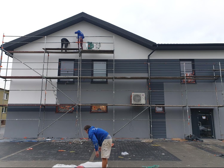 Dwukolorowa elewacja budynku w trakcie malowania. Widoczne rusztowania i pracownicy przy pracy. Fragment budynku w kolorze szarym i białym.