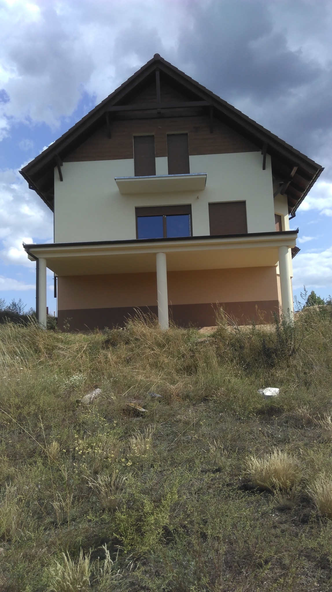 Dwukondygnacyjny dom z beżową elewacją i drewnianym dachem, widziany z dołu z zarośniętego wzgórza. Widoczne kolumny podtrzymujące balkon i brązowe okiennice.
