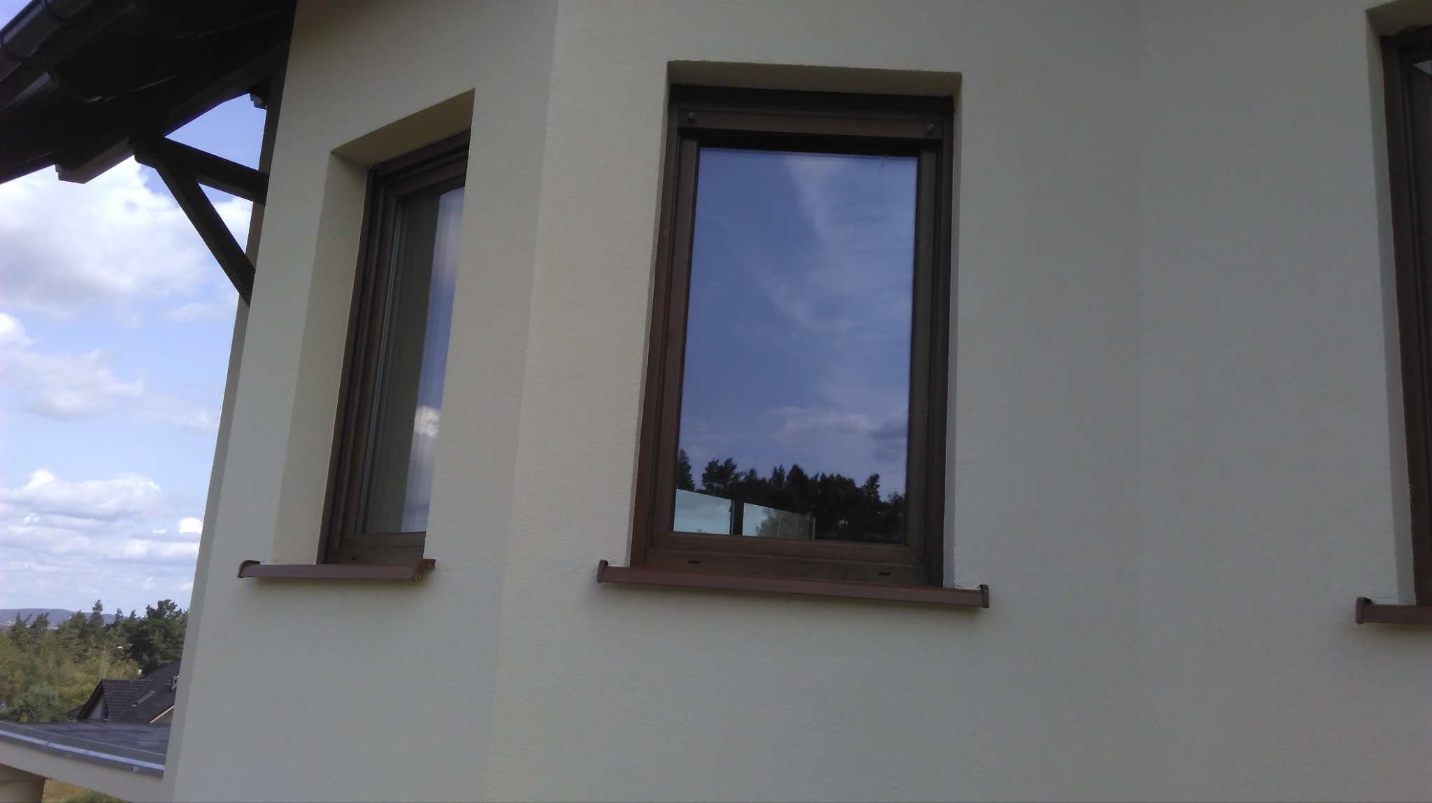 Elewacja domu po malowaniu w jasnym odcieniu, z brązowymi ramami okiennymi. Widoczne dwa okna z odbiciem nieba i drzew. Ujęcie z dołu, ukazujące fragment dachu.