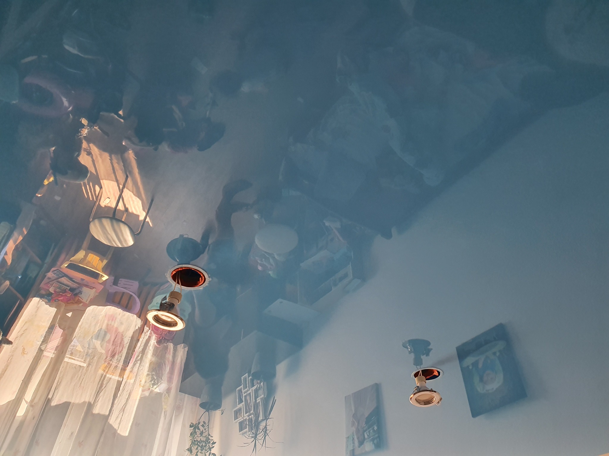 Błękitny sufit napinany z połyskiem lustrzanym, odbijający wyposażenie pokoju: lampy, zasłony, meble. Widoczne mocowania lamp sufitowych.