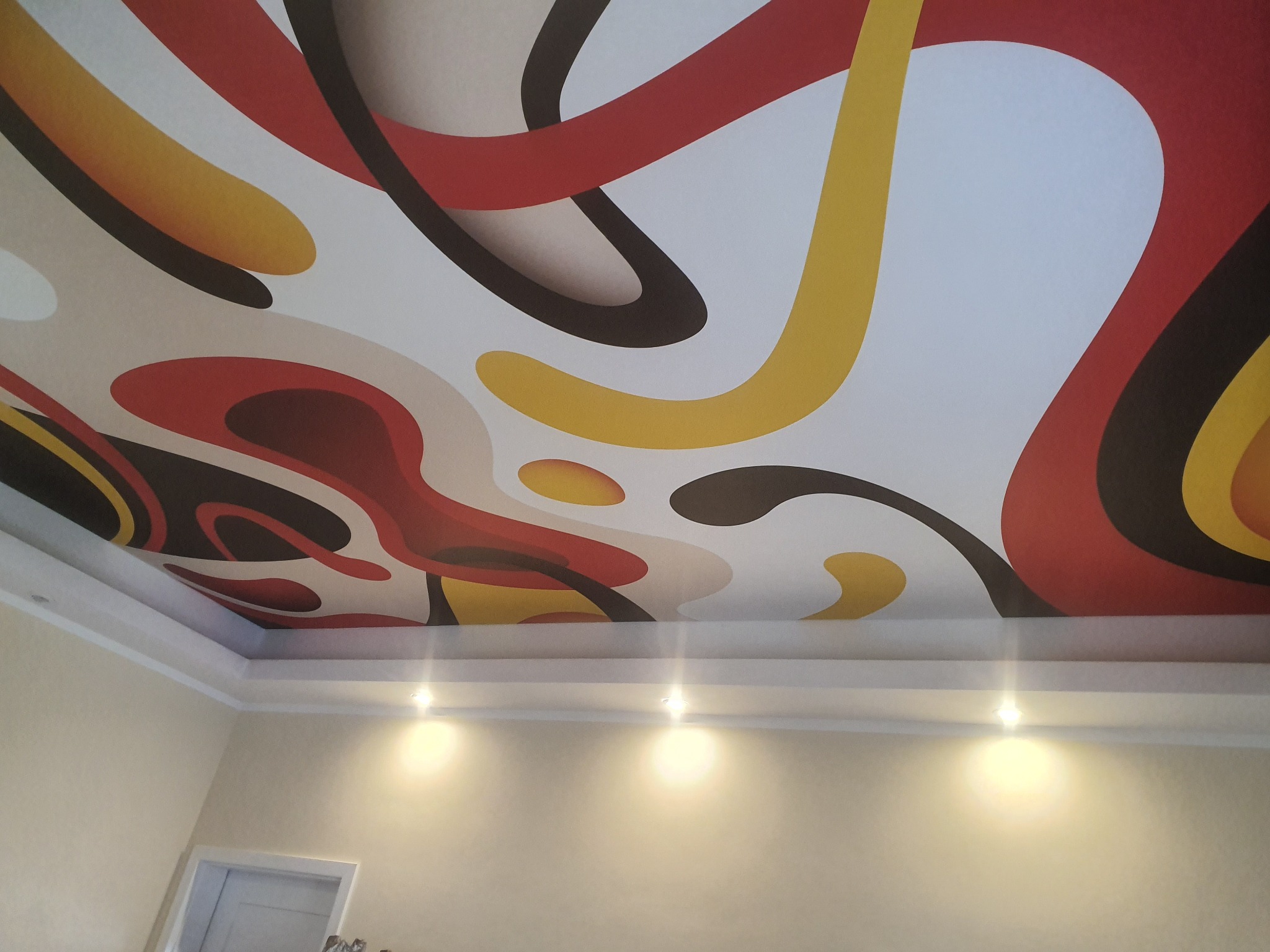 Sufit napinany z abstrakcyjnym wzorem w kolorach czerwonym, żółtym i czarnym. Widoczne oświetlenie punktowe i fragment ściany. Nietypowy design sufitu.