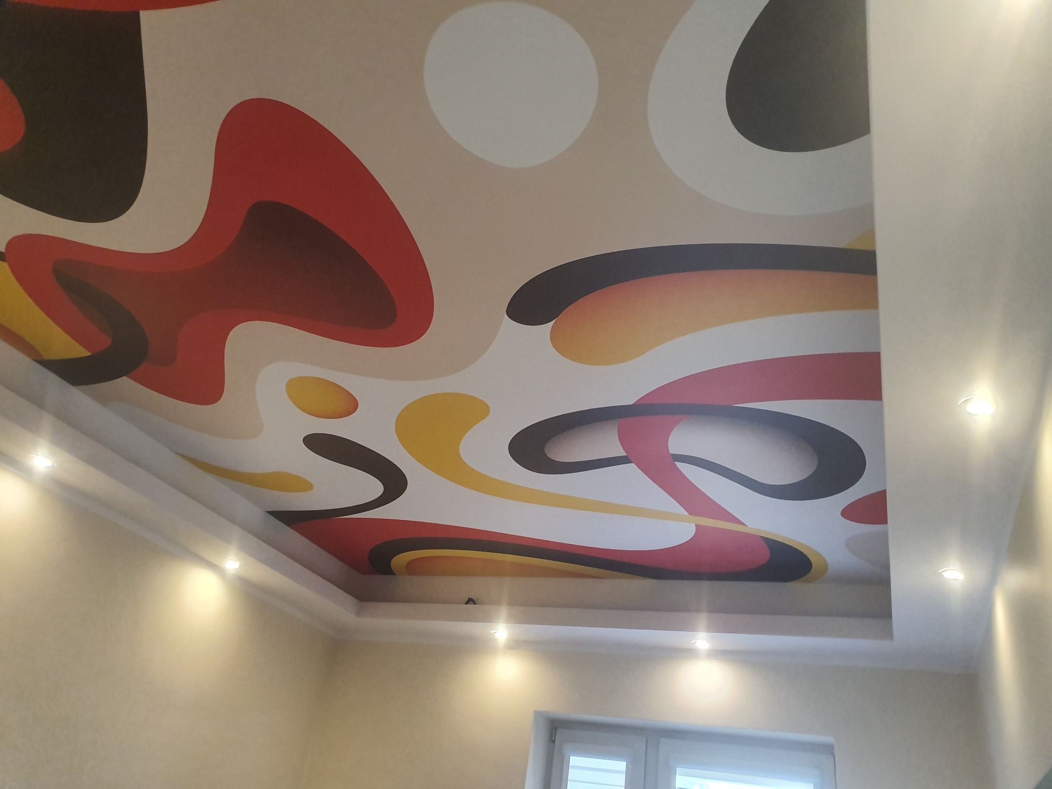 Sufit napinany z abstrakcyjnym wzorem w kolorach czerwonym, żółtym i czarnym. Oświetlenie punktowe podkreśla unikalny design. Widok z dołu.