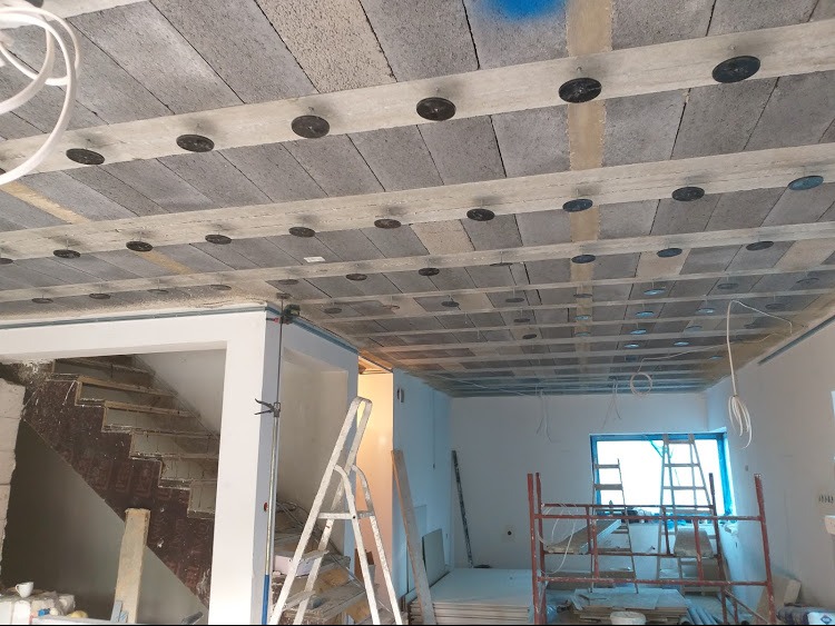 Sufit z zamocowanymi elementami izolacyjnymi, widoczne betonowe bloki i elementy mocujące. W tle pomieszczenie w trakcie remontu, drabiny i rusztowania.
