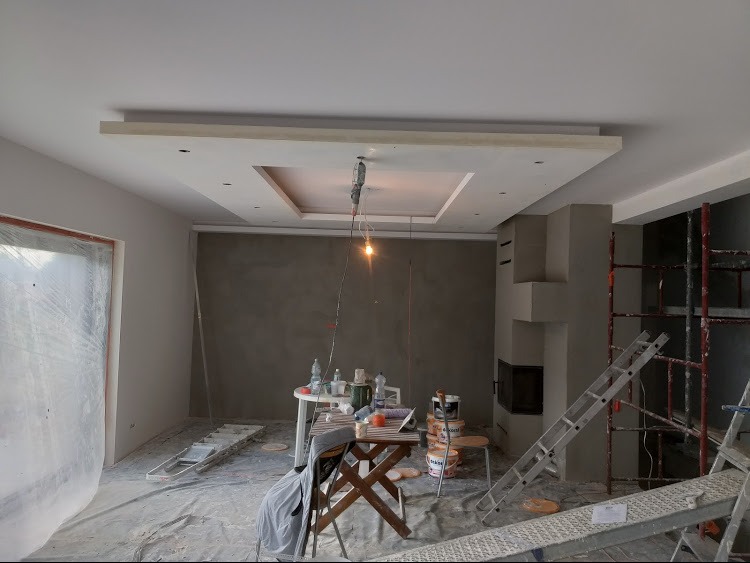 Wnętrze w trakcie prac wykończeniowych: szpachlowane ściany, zabudowa sufitu z oświetleniem, drabiny, narzędzia i materiały budowlane widoczne w pomieszczeniu.