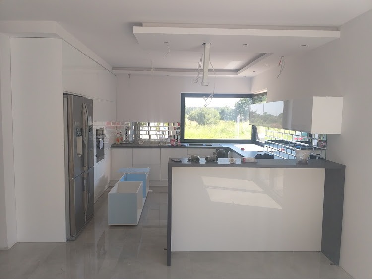 Nowoczesna kuchnia w trakcie montażu: białe szafki, srebrna lodówka, okno z widokiem na zieleń. Widoczne niedokończone elementy i narzędzia.