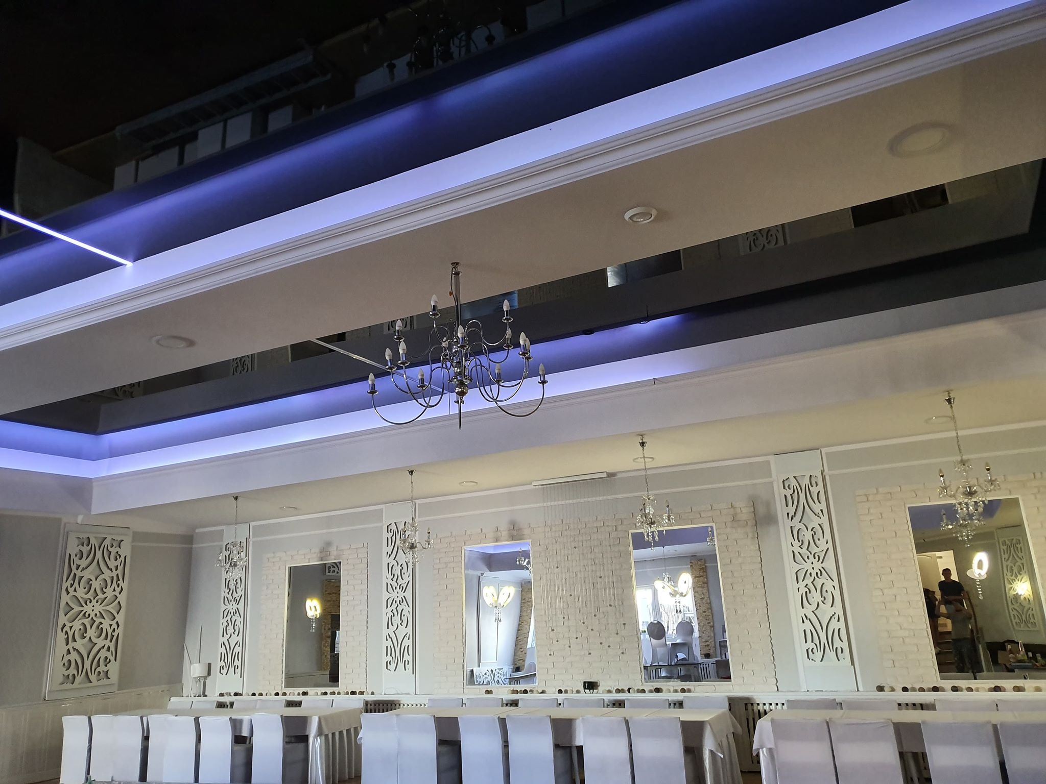 Elegancka sala bankietowa z dekoracyjnymi panelami ściennymi, lustrami i kryształowymi żyrandolami. Sufit podwieszany z niebieskim oświetleniem LED. Białe krzesła i stoły.