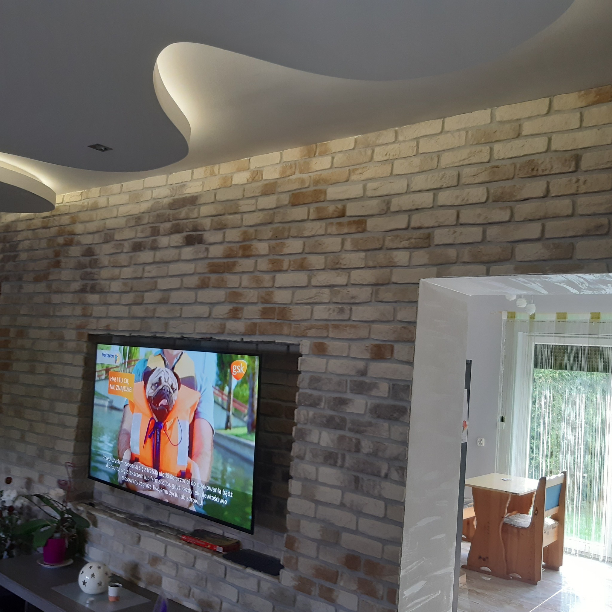 Salon z ceglaną ścianą, na której wisi telewizor. Sufit podwieszany z oświetleniem LED. Widoczne krzesła i stół w tle. Wnętrze po remoncie z dekoracyjnym wykończeniem.