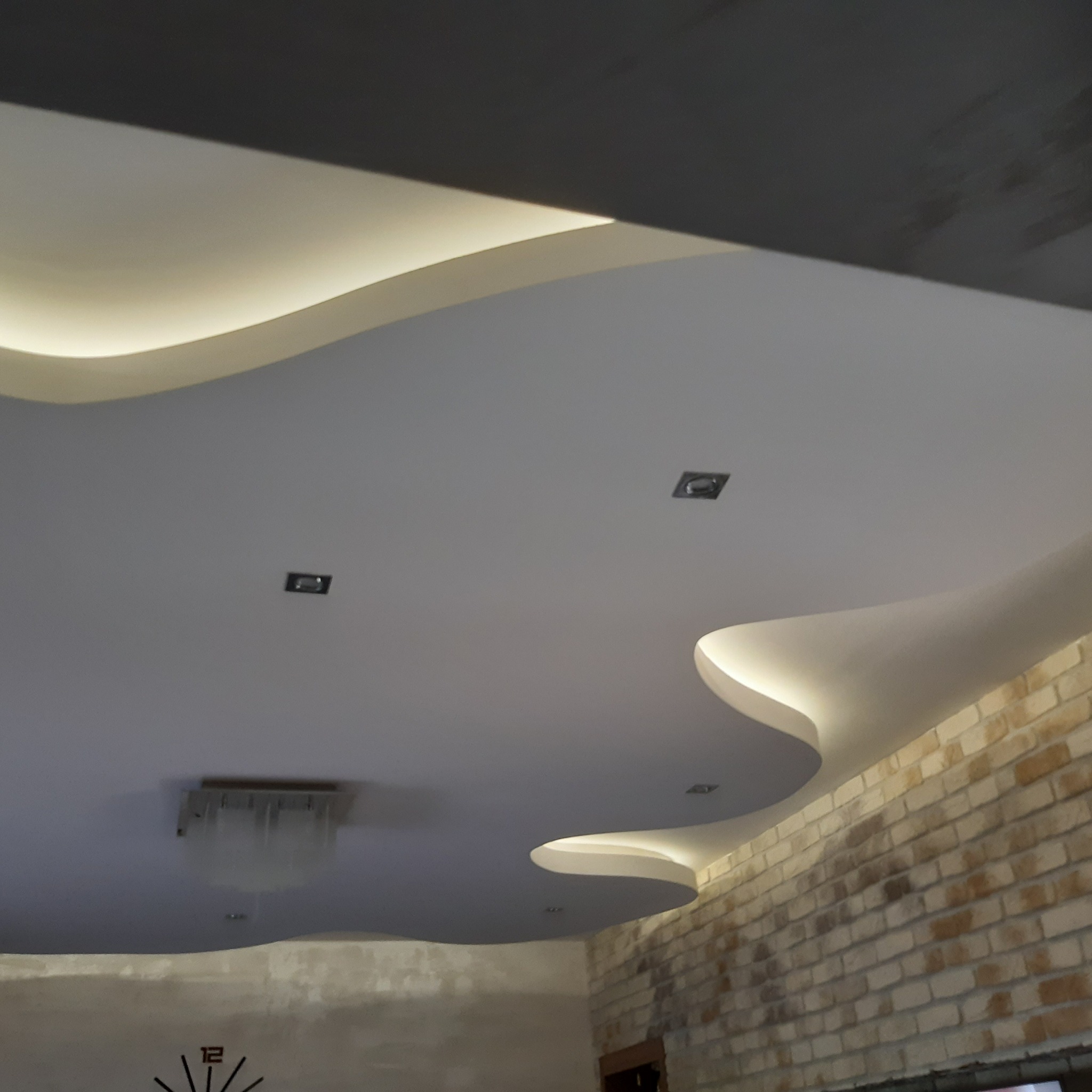 Falisty sufit podwieszany z oświetleniem LED i punktowymi lampami, połączony z ceglaną ścianą, tworząc unikalny design wnętrza.