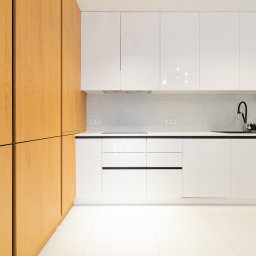 M3 Martin Paterak - Nowoczesna kuchnia z białymi szafkami na wysoki połysk i drewnianymi frontami. Minimalistyczny design, czarne akcenty i jasna podłoga. Oświetlenie LED pod szafkami.