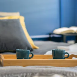 Dwie turkusowe filiżanki na drewnianej tacy na łóżku z szarą narzutą i dekoracyjnymi poduszkami. W tle rozłożona książka na stoliku nocnym.