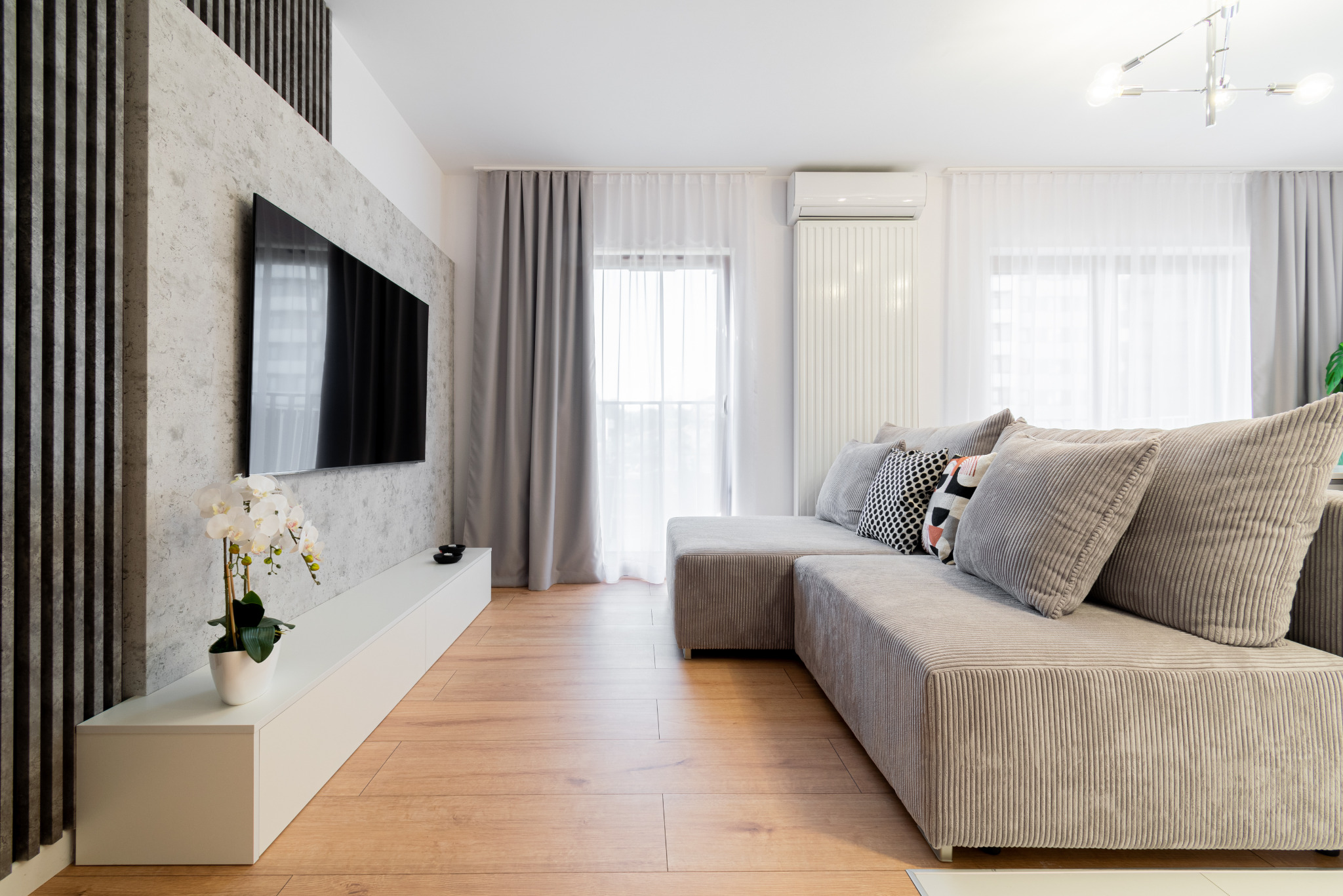 Jasne wnętrze salonu z szarą, narożną sofą z poduszkami, białymi zasłonami i niską szafką RTV z kwiatami. Na ścianie telewizor i dekoracyjne, czarne listwy.