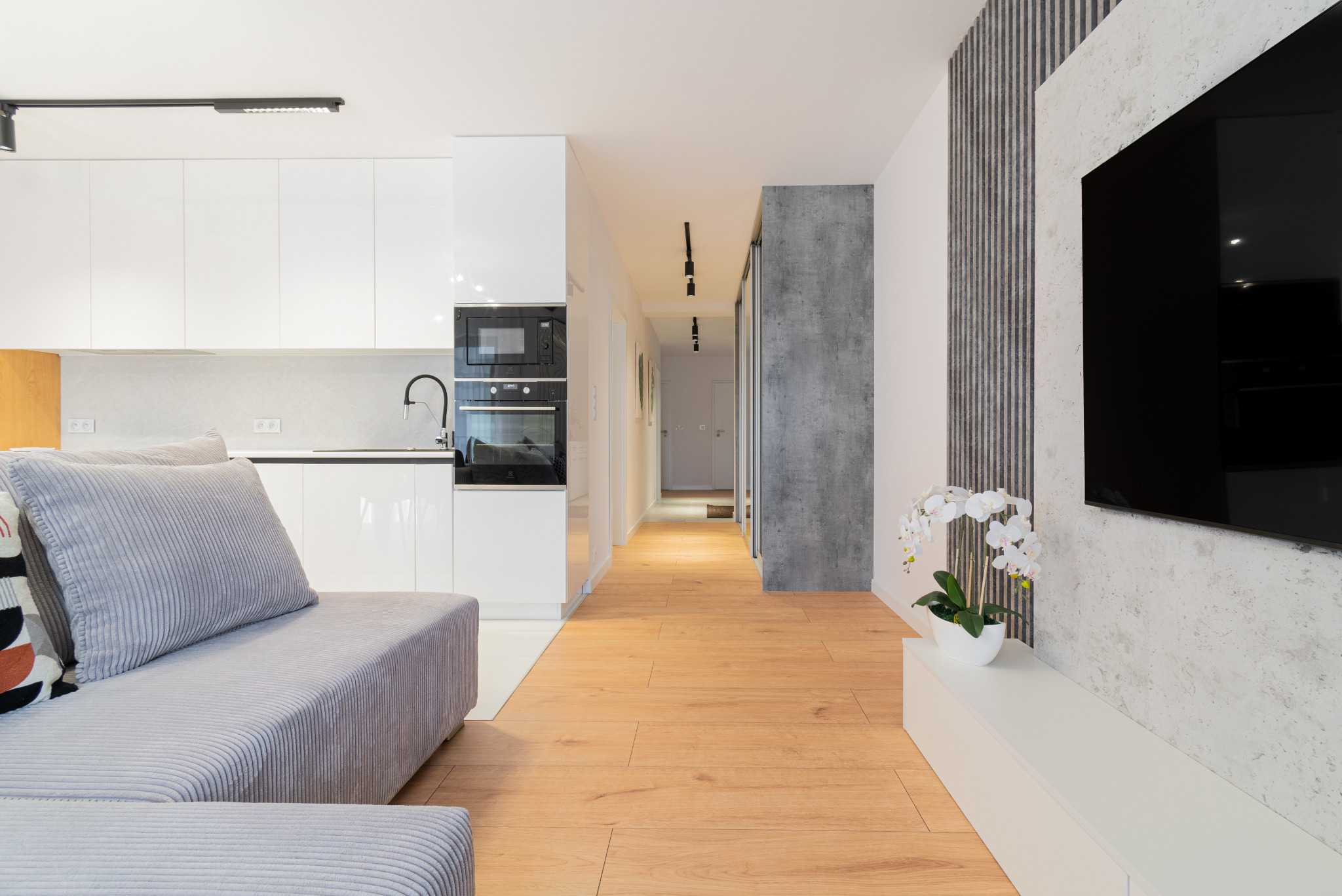 Nowoczesne wnętrze: jasna kuchnia z białymi szafkami, drewniana podłoga, szara sofa i telewizor na tle ściany z dekoracyjnym panelem. Minimalistyczny design.
