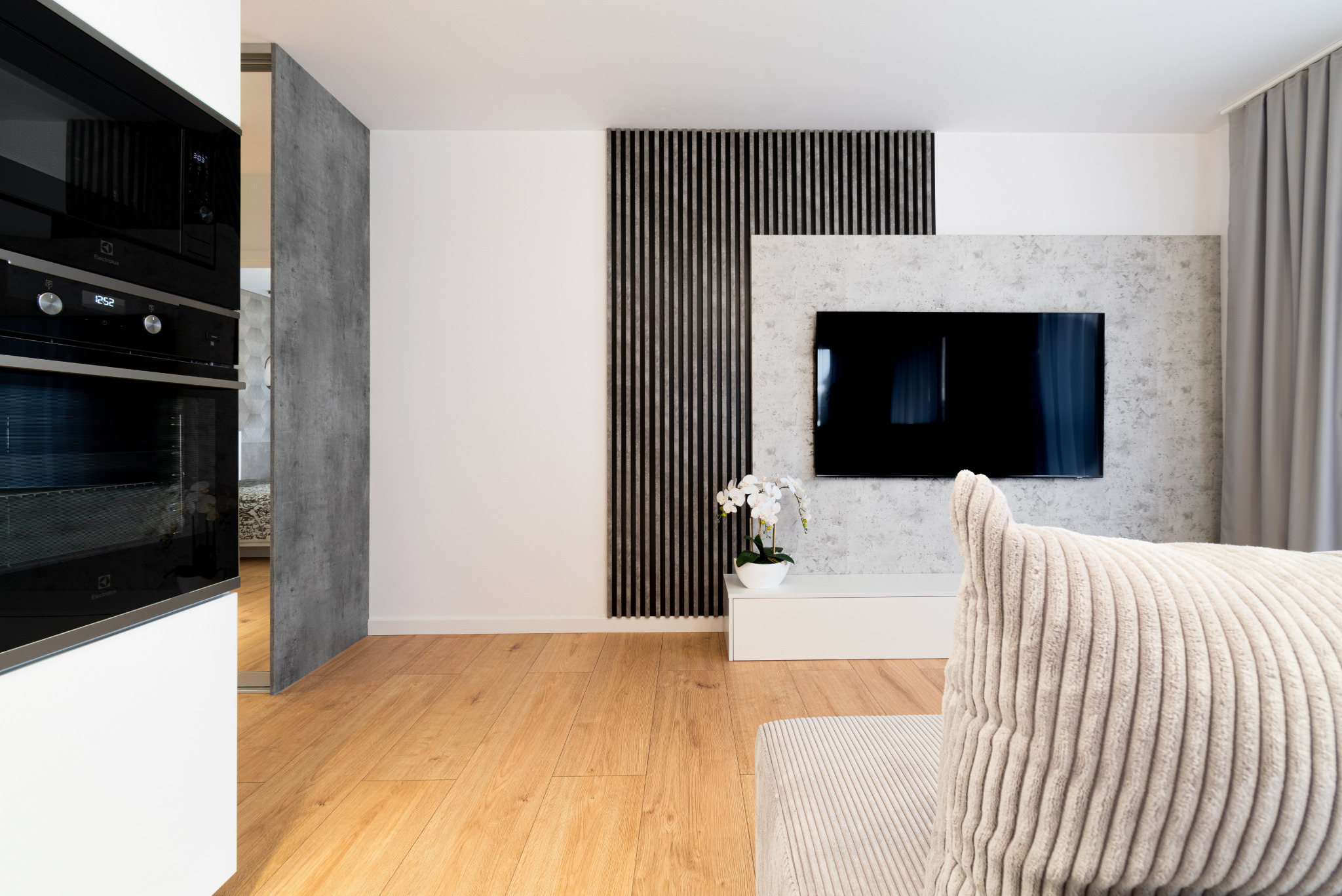 Jasne wnętrze salonu z drewnianą podłogą, szarą ścianą z telewizorem i dekoracyjnymi lamelami. W tle zabudowa kuchenna z piekarnikiem.