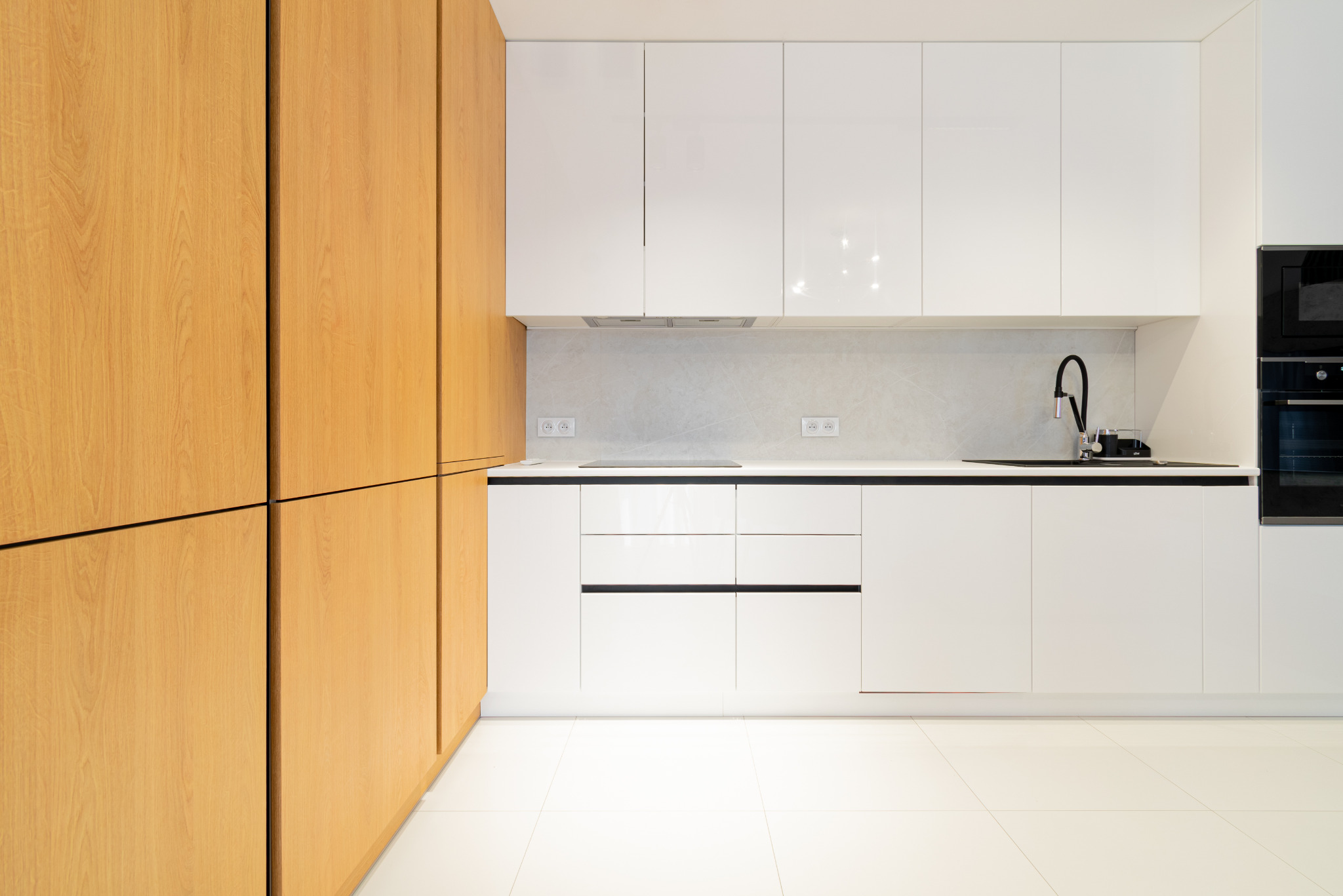 Nowoczesna kuchnia z białymi szafkami na wysoki połysk i drewnianymi frontami. Minimalistyczny design, czarne akcenty i jasna podłoga. Oświetlenie LED pod szafkami.