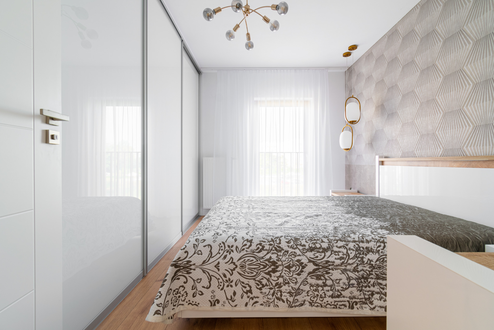 Nowoczesna sypialnia z łóżkiem w kwiatowy wzór, białą szafą przesuwną i geometryczną tapetą. Całość doświetlona przez stylowe lampy.