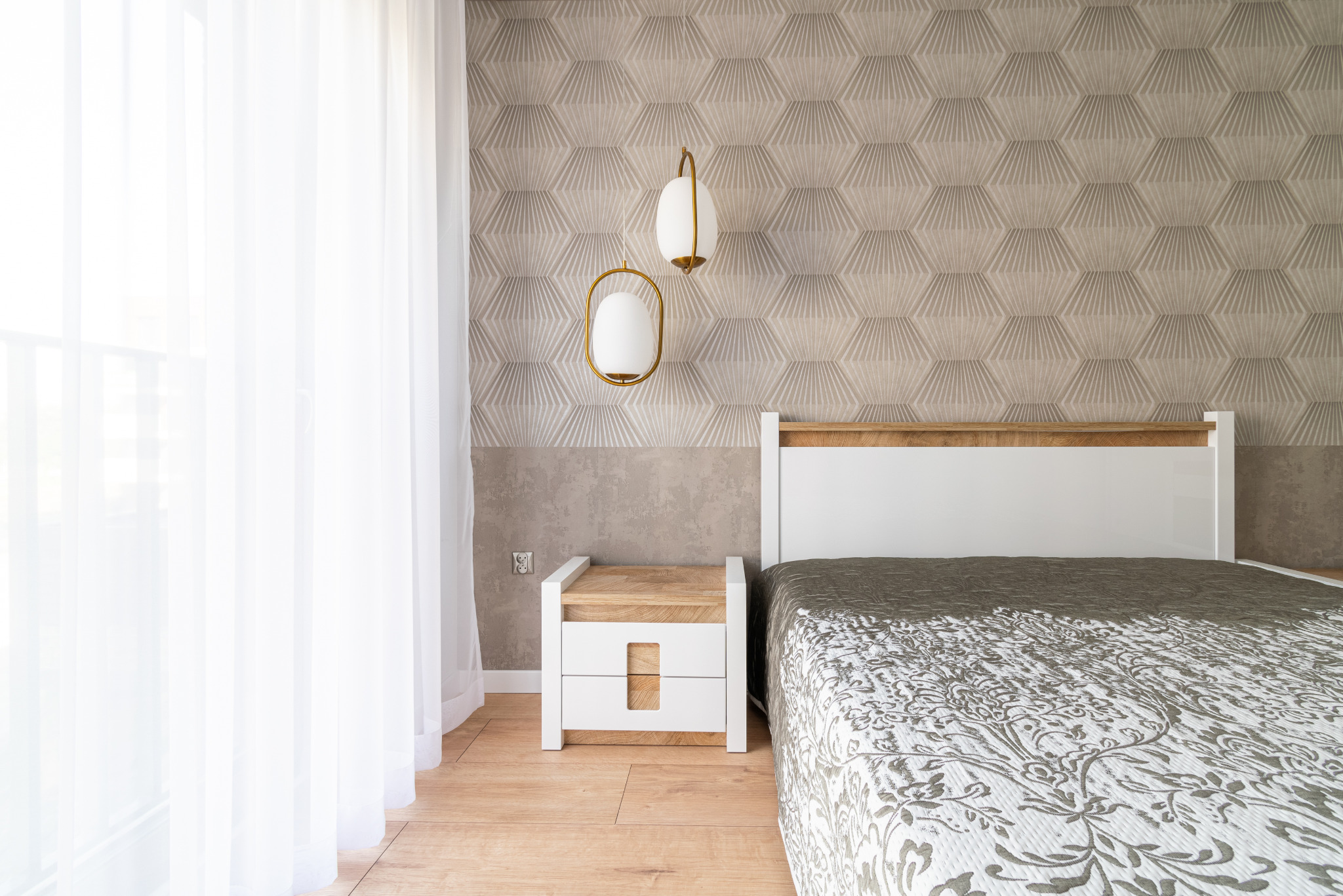 Jasna sypialnia z łóżkiem, szafką nocną i lampami wiszącymi na tle geometrycznej tapety. Delikatne światło wpadające przez okno z firanką.