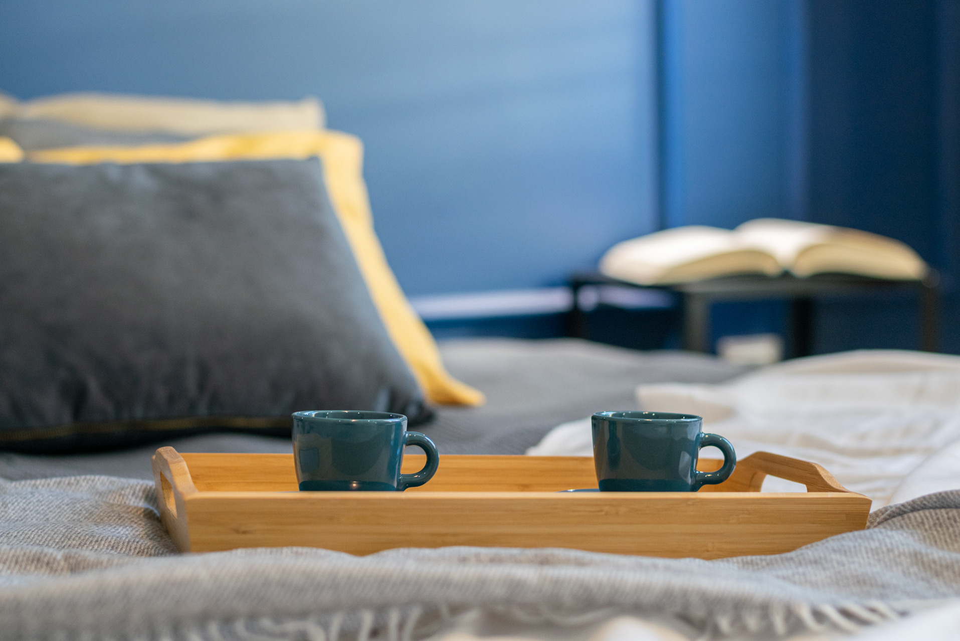 Dwie turkusowe filiżanki na drewnianej tacy na łóżku z szarą narzutą i dekoracyjnymi poduszkami. W tle rozłożona książka na stoliku nocnym.