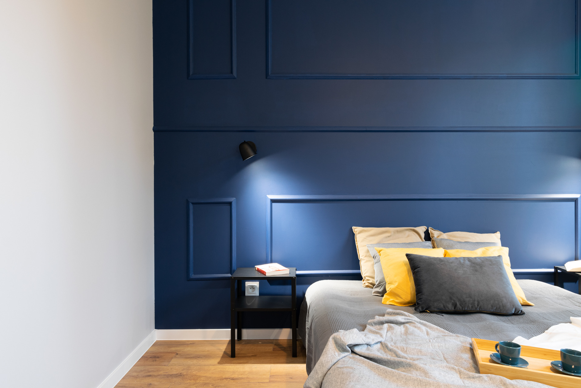 Stylowa sypialnia z łóżkiem, niebieską ścianą z dekoracyjnymi listwami i czarną lampką nocną. Na łóżku poduszki i taca z filiżankami. Minimalistyczny design.