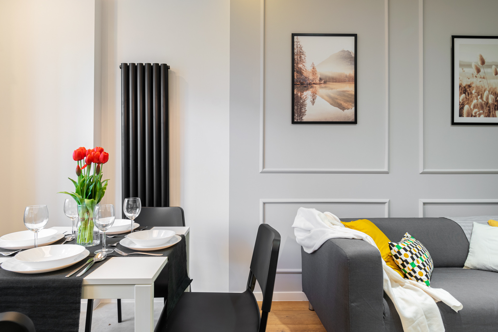 Elegancka jadalnia z minimalistycznym stołem, nakrytym do posiłku, z bukietem czerwonych tulipanów i szarą sofą w tle. Na ścianie wiszą obrazy.
