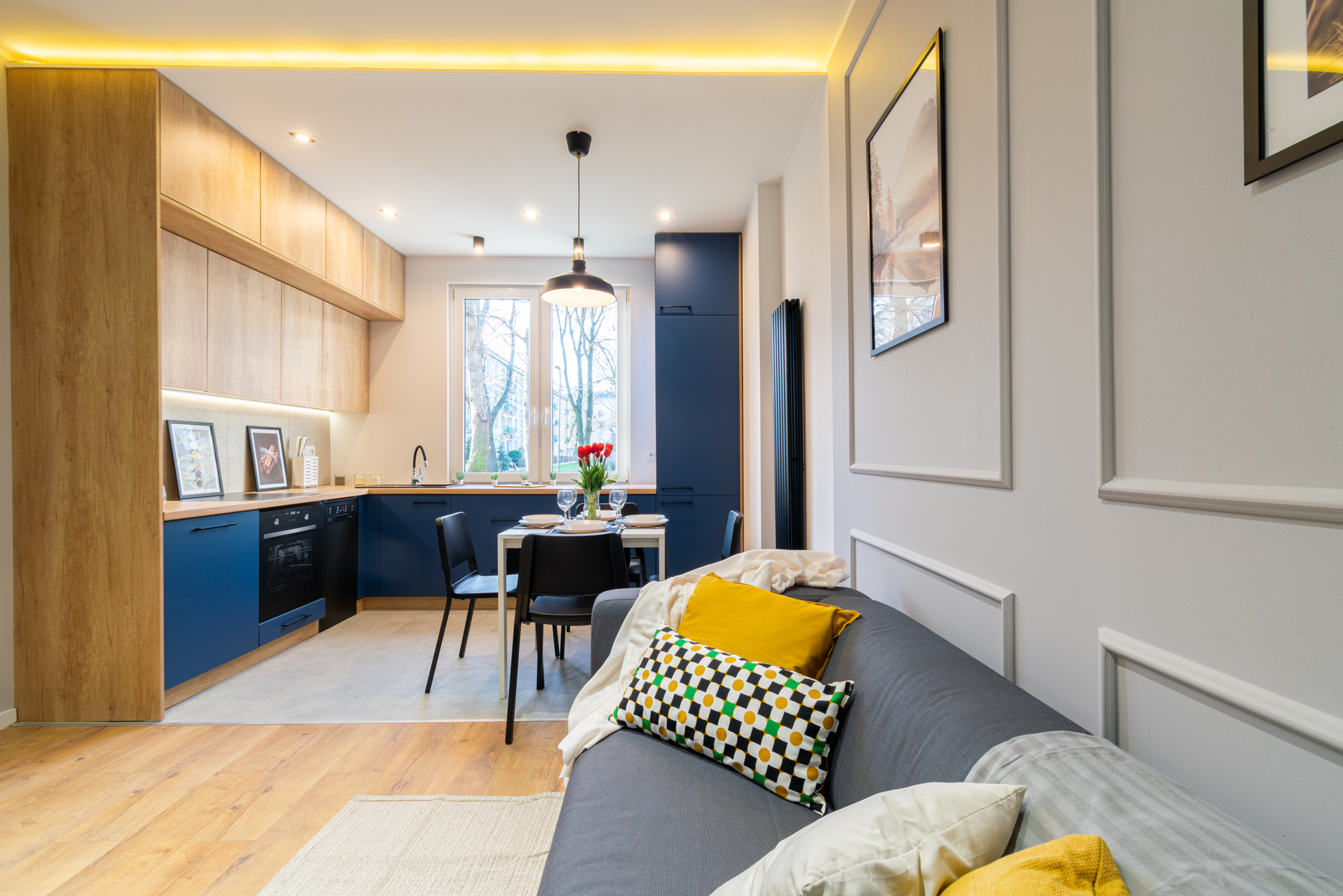 Nowoczesne wnętrze: granatowo-drewniana kuchnia z jadalnią i szarą sofą. Wazon z tulipanami na stole. Oświetlenie LED podszafkowe i sufitowe.