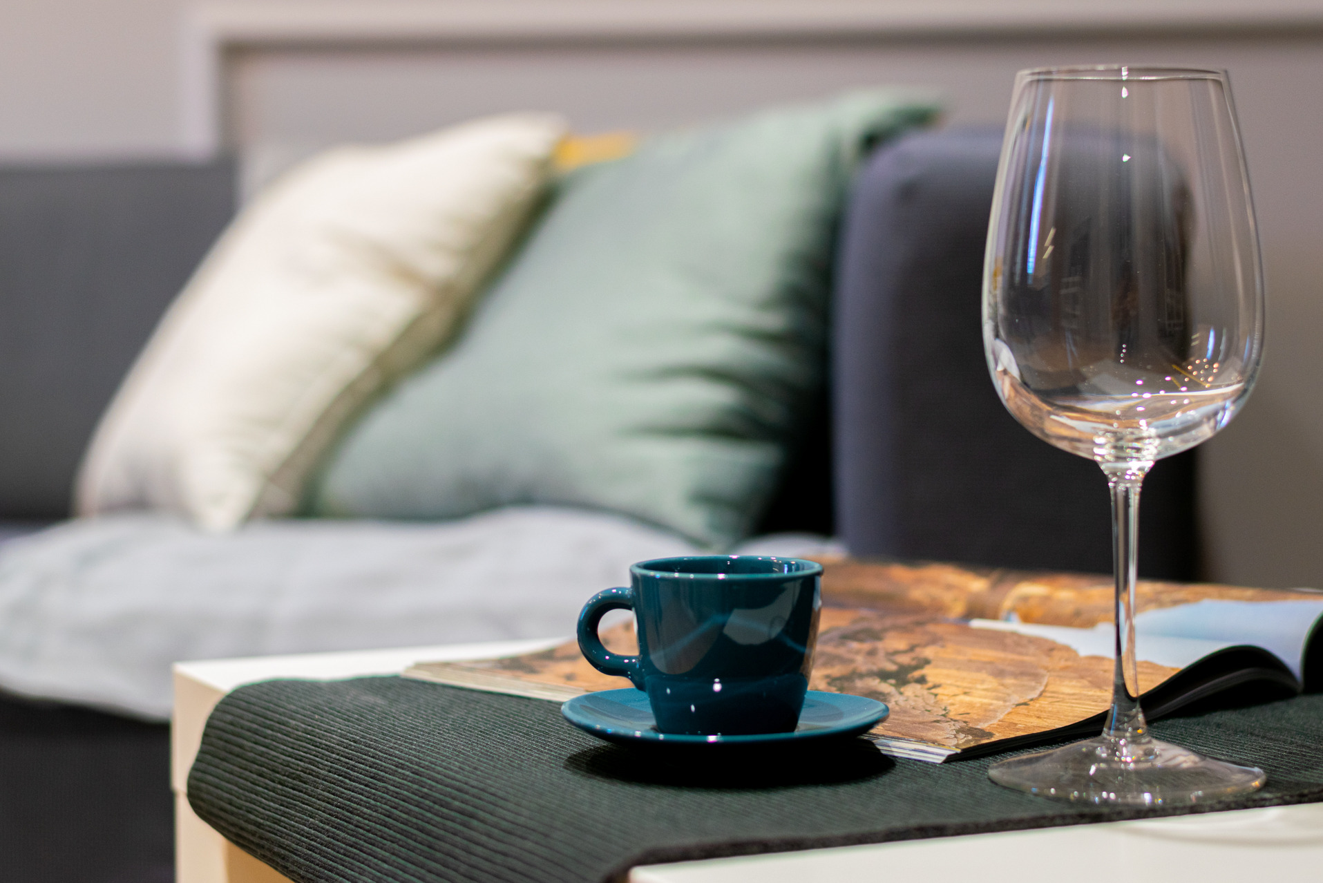 Turkusowa filiżanka espresso i kieliszek na stole z magazynem wnętrzarskim, w tle poduszki na szarej sofie. Fotografia wnętrza z bliska, miękkie oświetlenie.