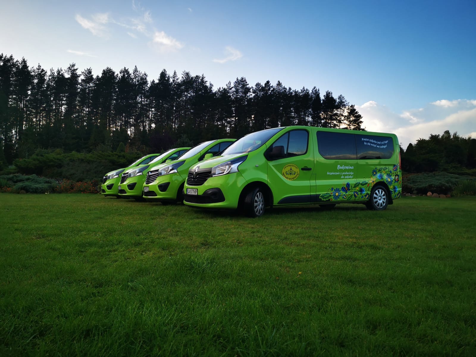Flota zielonych busów z dekoracyjnym motywem roślinnym, ustawiona rzędem na trawie na tle lasu i nieba. Widoczny napis 'Bukowina'.