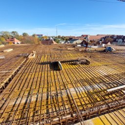 Tombud - Zbrojenie stropu budynku w trakcie budowy, widoczne żółte szalunki i stalowe pręty. W tle zabudowa miejska i błękitne niebo.