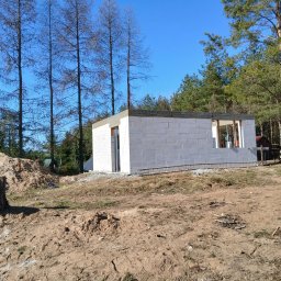 Tombud - Wznoszenie ścian parterowego domu z betonu komórkowego na fundamencie, widoczne zbrojenie i otoczenie budowy z drzewami w tle.