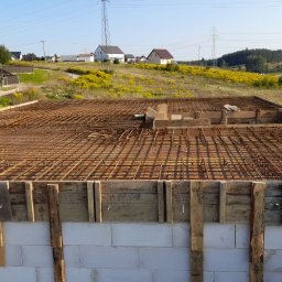 Tombud - Zbrojenie płyty fundamentowej na budowie domu jednorodzinnego. Widoczne białe bloczki fundamentowe, drewniane szalunki i siatka zbrojeniowa. W tle zabudowa wiejska.